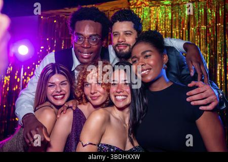 Eine Gruppe fröhlicher, eleganter Freunde posiert gemeinsam für ein Foto bei einer Nachtclub-Party mit Disco-Lichtern und goldenem Lametta-Vorhang Stockfoto