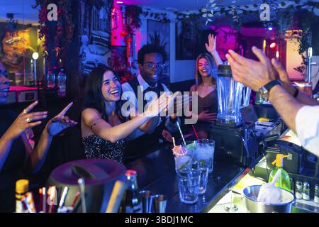 Fröhliche Multi-ethnische Freunde klatschen und genießen lebhafte Cocktails, teilen Lachen und Aufregung in einer lebhaften Nachtlebenatmosphäre Stockfoto