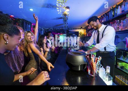 Fröhliche Gruppe von Freunden tanzen und Cocktails in einer Nachtclub-Bar, während ein Barkeeper Getränke zubereitet Stockfoto