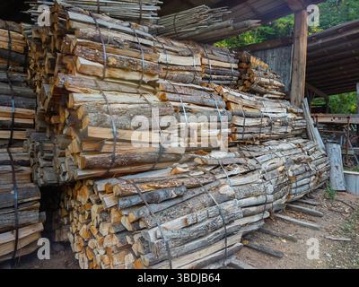 Mit Metallbändern verbundene, stapelbare Brennholzbündel werden unter einem hölzernen Unterstand in einem Außenbereich gelagert. Gehackte Baumstämme zeigen Rindenstruktur und sind vielfältig Stockfoto