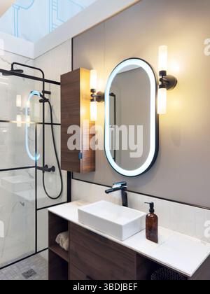 Modernes Badezimmer mit rechteckigem Waschbecken, beleuchtetem ovalem Spiegel, Holzwaschtisch mit integriertem Stauraum und Glasdusche mit B Stockfoto