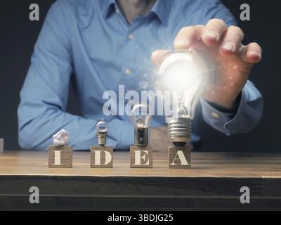 Vier Holzblöcke mit der Aufschrift New Idea und einer zerknitterten Papierkugel und drei unterschiedlich großen Glühbirnen, neue Ideen und Kreativitätskonzept Stockfoto