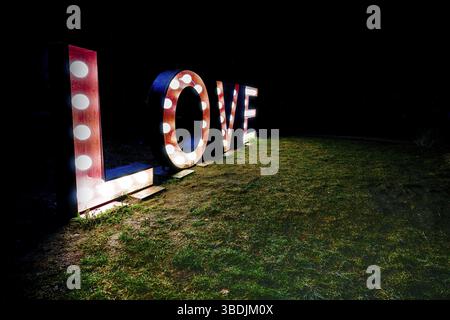 Das Wort Liebe geschrieben in hell erleuchteten Buchstaben in der Nacht mit roten und weißen Glühlampen auf einer Wiese in einem dunklen schwarzen Nacht Stockfoto