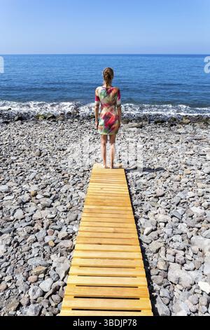 Frau, die auf einem Holzweg am steinigen Strand und Meer steht Stockfoto