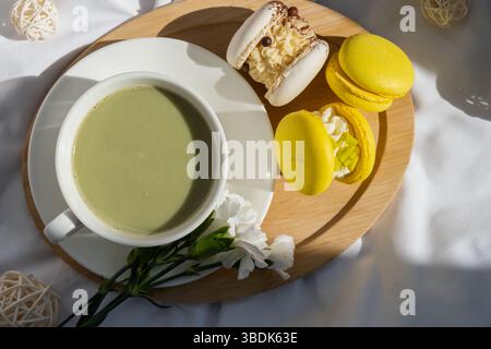 Süßes gelbes Macaron Dessert Tasse grüner Matcha Tee Latte auf Pflanzenbasis in gemütlicher Morgenliegenatmosphäre. Kreative Urlaubsidee mit romantischem Date. Pflanzliche präbiotische Faser Superfood Getränk Antioxidans-reiche Immunität-Boosting Kaffee Alternativen Getränk Stockfoto