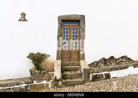 Santa Maria de Marvao, Portugal - 30. März 2022: Weiß getünchte Hausfront mit alter Holztür in Steinrahmen auf einer gepflasterten Straße Stockfoto