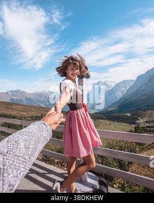 Schöne junge Frau, die traditionelle bayerische Kleidung trägt - Dirndl oder tracht, während sie die Hand der Männer hinter der Kamera vor dem Fernglas hält. Stockfoto