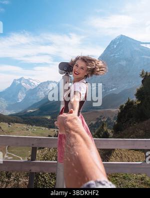 Schöne Frau in traditioneller bayerischer Kleidung - Dirndl oder tracht - beobachtet die Landschaft der europäischen Alpen durch ein Fernglas in der Schweiz Stockfoto
