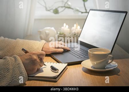 Hände einer älteren Frau, die mit dem Laptop ein günstiges Online-Shopping für Weihnachtsgeschenke nutzt und Notizen auf einem Tisch mit Adventskerzen, Schweinebank und macht Stockfoto