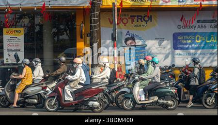 DANANG, VIETNAM, 25. April 2025, urbane Szene mit Menschen auf Motorrollern, die bei Tageslicht an einer geschäftigen Kreuzung warten, umgeben von farbenfrohen storef Stockfoto