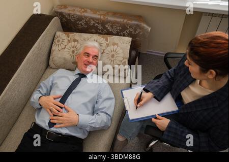 Ein älterer Mann liegt während eines Termins mit einem Psychotherapeuten auf einer Couch. Ein älterer männlicher Patient in einer individuellen Konsultation mit einer Frau Stockfoto