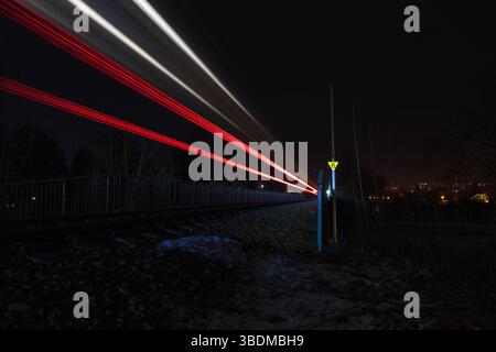 Lichtspuren eines Zuges, der nachts auf der Eisenbahnstrecke über die Brücke in die Stadt gefahren wird. Leuchtende rote und weiße Lichter symbolisieren Geschwindigkeit, Technologie und Stockfoto