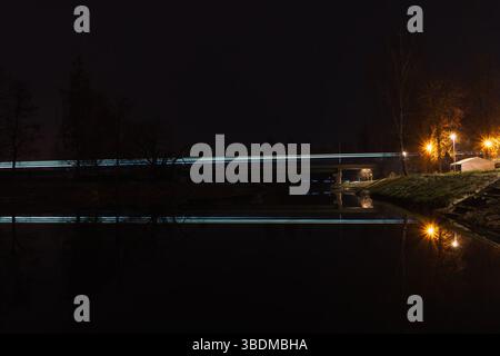Lichtspuren eines Zuges in der Nacht auf einer Eisenbahnstrecke, die über eine Brücke mit Reflexion in der Moldau fährt. Leuchtende weiße Lichter symbolisieren Geschwindigkeit, Technol Stockfoto