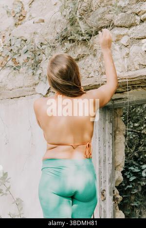 Frau, zurück, Wand - Frau mit langen braunen Haaren, die ein grün-weißes Tie-Dye-Top und grüne Leggings trägt, steht vor einer weißen Steinmauer. Stockfoto