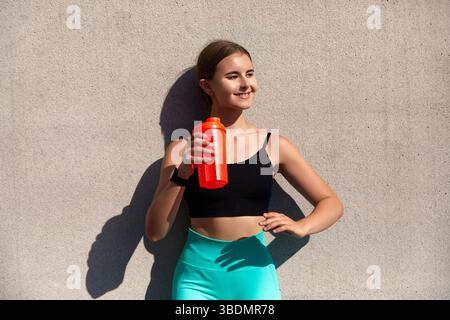 Eine junge Frau in stilvoller Aktivkleidung posiert mit einer leuchtend roten Wasserflasche an einer Betonwand und betont so das Engagement für Fitness und Hydration Stockfoto