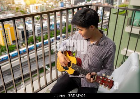 Asiatischer junger Mann, der eine Akustikgitarre in seinem persönlichen Wohnzimmer hält und spielt. Stockfoto