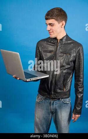 Junger Mann mit schwarzer Lederjacke, der einen Laptop hält und auf blauem Hintergrund lächelt Stockfoto