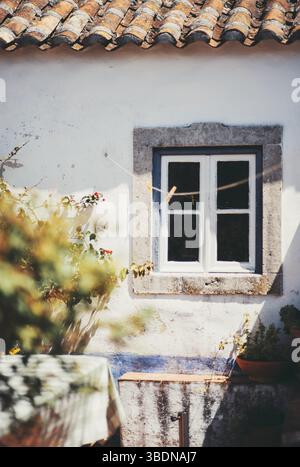 Rustikale weiß getünchte Wand mit einem kleinen Holzfenster, das in Stein eingerahmt ist, unter einem Dach aus sonnenbeleuchteten Terrakotta-Fliesen, umgeben von Kletterpflanzen und einem gemütlichen Ou Stockfoto