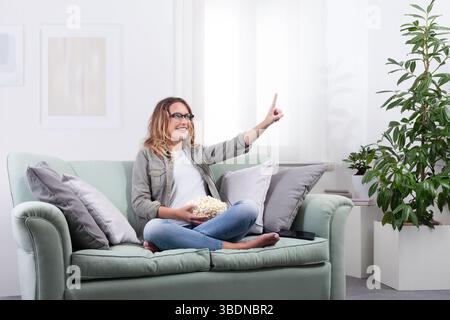 Frau mit einer intelligenten Brille, Popcorn in der Hand. Sie zeigt mit dem Finger nach vorne, als ob sie mit einem virtuellen Display interagiere. Blick in den Weltraum und geniessen digi Stockfoto