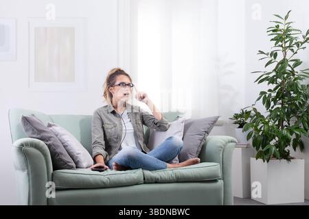 Staunte junge Frau, die eine intelligente Brille mit Smartphone neben sich trägt. Sie berührt ihre Brille und sieht nach vorne, als ob sie mit digitalen Inhalten interagieren würde Stockfoto