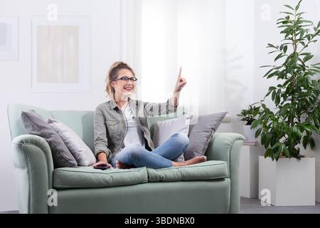 Lächelnde junge Frau mit einer intelligenten Brille, die zu Hause auf der Couch sitzt und neben ihr ihr ihr Smartphone hat. Sie blickt nach vorne und Touchscreen, interagiert mit di Stockfoto