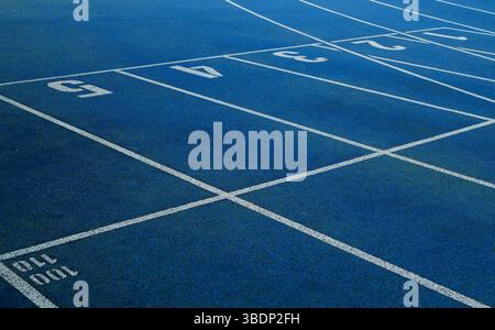 Geschwungene blaue Laufstrecke mit weißen Spurmarkierungen in einem Sportstadion im Freien. Grünes Gras und städtische Wohngebäude im Hintergrund. Stockfoto