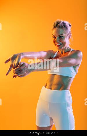 Schöne reife sportliche Frau mit Sportbekleidung beim Training – fit, reife Frau in sportlicher Kleidung, die vor einem leuchtenden orangen Hintergrund trainiert, s Stockfoto