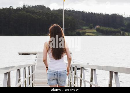 Junge Frau mit langen Haaren von hinten gesehen, auf einem Dock spazieren und auf einen ruhigen See blicken, der von bewaldeten Hügeln umgeben ist Stockfoto