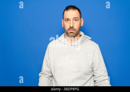Ein hispanischer Mann mit Bart in seinen 40ern, der ein graues Sweatshirt trägt, blickt mit einem ernsten und besorgten Ausdruck geradeaus, isoliert in einem blauen Studio Stockfoto