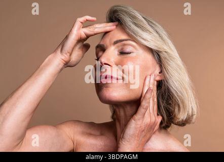 Schönheitsprofil der schönen Reifen Frau, Konzepte über Kosmetik, Hautpflege und Wellness. Reife Frau mit geschlossenen Augen, die sanft das Gesicht berührt, verkörpert Stockfoto
