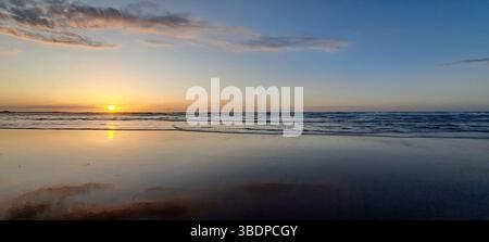 Der ruhige Sonnenaufgang färbt den Himmel mit Gold und reflektiert sich auf dem nassen Sand eines friedlichen Strandes, während sanfte Wellen hereinbrechen Stockfoto