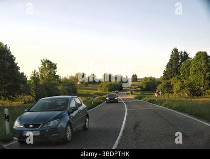 Das Bild zeigt eine Landstraße mit mehreren Autos, die in beide Richtungen fahren. Die Straße führt sanft durch eine grüne Landschaft mit Bäumen und Gras auf bo Stockfoto