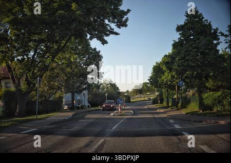 Das Bild zeigt eine ruhige Wohnstraße mit einem Kreisverkehr in der Mitte. Auf beiden Seiten der Straße säumen Bäume, die Schatten spenden. Eine rote ca. Stockfoto