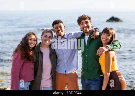 Gruppe fröhlicher junger Freunde, die einen sonnigen Tag am Strand genießen, lächeln und das Meer im Hintergrund genießen. Stockfoto