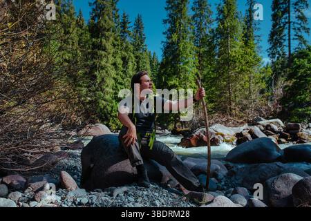 Junger Mann Wanderer, der auf einem großen Stein in einer Bergschlucht in der Nähe des Flusses sitzt und wegblickt Stockfoto