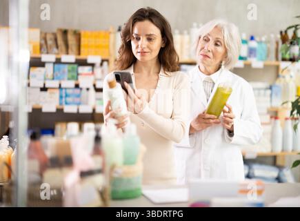 Frau in der Apotheke scannt QR-Code auf Shampoo, ältere Frau Apothekerin im Hintergrund Stockfoto