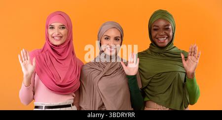 Drei Muslimische Damen Winken Hallo Geste Über Gelbem Hintergrund, Panorama Stockfoto