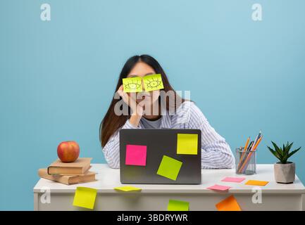 Erschöpfter Schüler. Porträt einer überarbeiteten Dame am Schreibtisch mit Aufklebern auf den Augen und mit Laptop, blauer Hintergrund Stockfoto