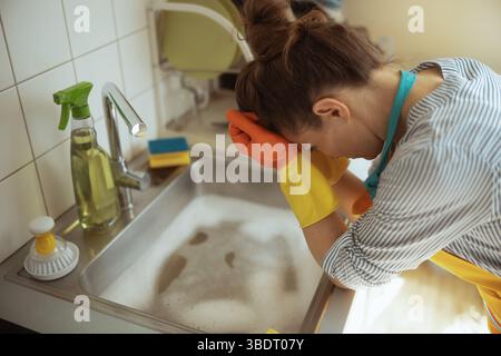 Eine Frau in Handschuhen lehnt sich mit dem Kopf nach unten auf das Spülbecken, erschöpft nach einem verstopften Abfluss und stehendem Seifenwasser. Stockfoto