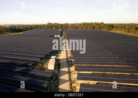 Ein großer Solarpark mit Reihen von Solarpaneelen erstreckt sich über die Landschaft. Schmaler Schotterweg verläuft durch die Mitte mit Versorgungsstrukturen in der Nähe. Dichter Wald grenzt an Panels im Hintergrund Stockfoto