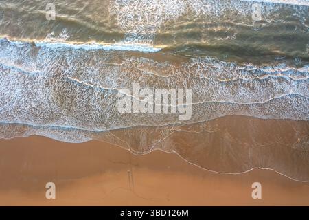 Drohnenaufnahme von oben nach unten mit Meereswellen, die auf den Sandstrand stürzen. Weiße Schaummuster bilden sich, wenn Wasser auf das Ufer trifft. Sand erscheint glatt mit leichten Markierungen in der Nähe der Wasserlinie Stockfoto
