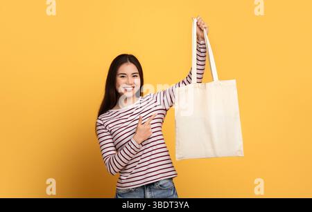 Die junge Frau posiert fröhlich mit einer wiederverwendbaren Tragetasche vor gelbem Hintergrund Stockfoto