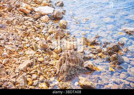 Felsige Küste mit verschiedenen Größen und Formen von Steinen, die sich in klares, flaches Wasser ausdehnen. Trockene Felsen am Ufer und die untergetauchten Steine im Wasser Stockfoto