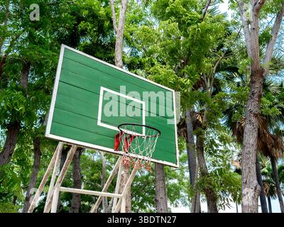 Alter leerer Basketballkorb aus grünen Holzpaneelen mit Netz im Garten mit hohen Bäumen in der Nähe des Strandes auf blauem Sommerhimmel Hintergrund. Stockfoto