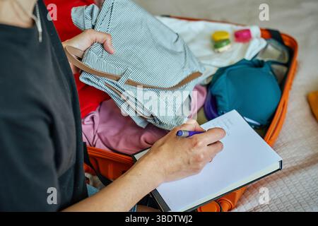 Frau bereitet sich auf die Reise vor, packt Koffer mit Kleidung und überprüft die Reisepackliste. Konzept der Urlaubs- und Urlaubsplanung Stockfoto