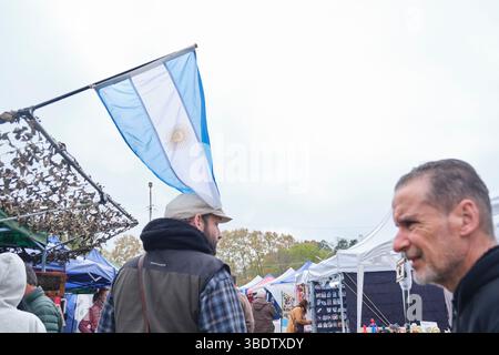 25. Mai 2025; Lobos, Buenos Aires, Argentinien: Argentinische Nationalflagge und Menschen, die durch die Verkaufsstände gehen, um Kunsthandwerk zu verkaufen Stockfoto