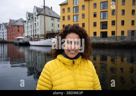 Porträt einer jungen brünetten Frau, die in eine gelbe Daunenjacke eingehüllt ist, mit dem Brosundet-Kanal in Alesund, Norwegen, hinter ihr, mit Stockfoto