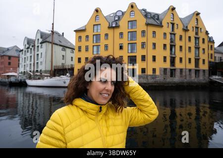 Porträt einer jungen brünetten Frau, die wegschaut und ihr Haar berührt, einen gelben Anorak trägt, mit dem Brosundet-Kanal in Alesund, Norwegen, hinter ihr, Witz Stockfoto