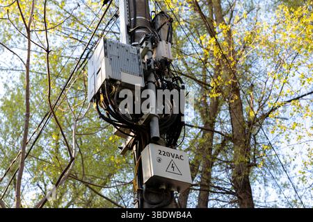 Elektro- und Kommunikationsgeräte, die an einem Versorgungsmast mit sichtbaren Kabeln und einem 220-VAC-Warnkasten montiert sind, umgeben von Bäumen Stockfoto