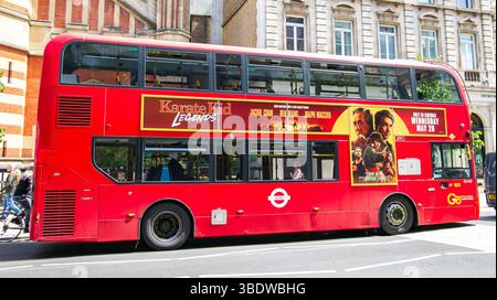 Roter Doppeldeckerbus mit Filmwerbung, der durch eine sonnige Stadtstraße in der Nähe historischer Gebäude in London fährt Stockfoto